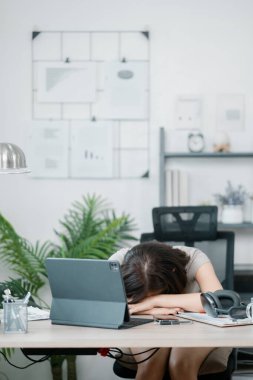 Çağdaş bir ofis ortamında başını masaya yaslayan yorgun bir kadın. Etrafı iş malzemeleri ve dizüstü bilgisayarla çevrili..