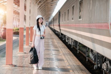 Sırt çantası ve kamerası olan genç bir kadın gezgin tren istasyonunda duruyor, yolculuğuna hazır..
