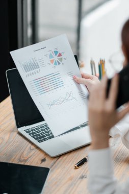Bir iş kadını, modern bir ofis ortamında dizüstü bilgisayar üzerinde çalışırken çeşitli grafikler ve çizelgeler içeren bir mali raporu inceliyor..