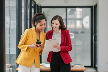 Modern bir ofiste işbirliği yapan, belgelerini inceleyen ve akıllı telefon kullanan şık ceketli iki profesyonel iş kadını..