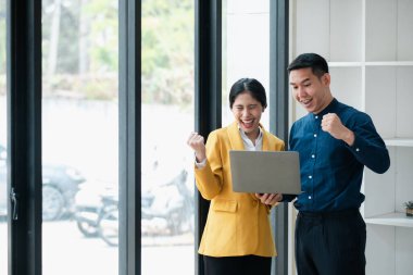 Modern ofis ortamında geniş pencereli bir dizüstü bilgisayara bakarken başarıyı kutlayan iki iş arkadaşı..