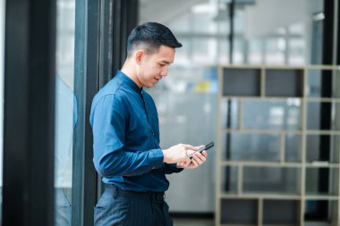 Mavi gömlekli profesyonel bir adam pencerenin yanında duruyor ve modern ofis ortamında akıllı telefonunu kullanıyor..