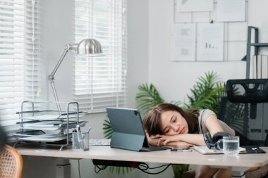 Genç bir kadın, bir dizüstü bilgisayar, belgeler ve ofis malzemeleriyle çevrili modern bir ofiste, bir masanın üzerinde dinleniyor..