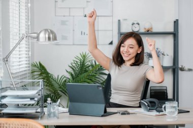 Kutlama için kollarını kaldıran neşeli genç bir kadın, modern bir ofiste bilgisayar ve belgelerle çalışıyor..