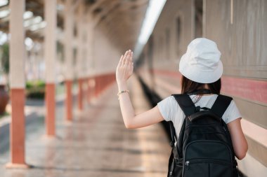 Şapka ve sırt çantası takan genç bir gezgin tren istasyonunda bir maceraya hazır bir şekilde el sallıyor..