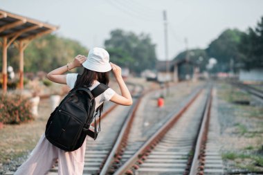 Sırt çantası ve şapka takan genç bir kadın tren raylarında duruyor. Macera ve keşfi simgeliyor. Seyahat ve açık hava temaları için mükemmel.