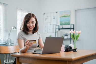 Genç bir kadın, dekorasyon ve doğal ışık ile çevrili modern bir ev ofisinde dizüstü bilgisayarla online alışveriş yapmak için kredi kartı kullanıyor..