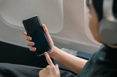 Uçakta kulaklık takarken akıllı telefon kullanan, modern seyahat ve uçuş eğlencesi sunan birinin yakın plan fotoğrafı..