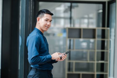 Modern ofis ortamında dururken akıllı telefon kullanan mavi gömlekli genç bir profesyonel..