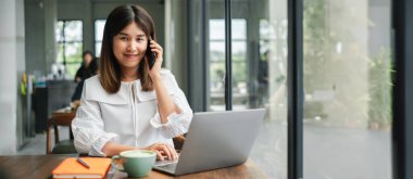 Dizüstü bilgisayarıyla çalışan ve modern ofis ortamında telefonla konuşan profesyonel bir kadın..