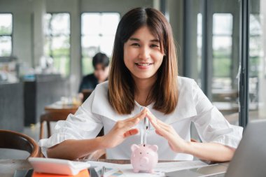 Parlak, modern bir ofis ortamında kumbarayı tutarken gülümseyen genç kadın, mali planlama ve tasarruf simgesi..