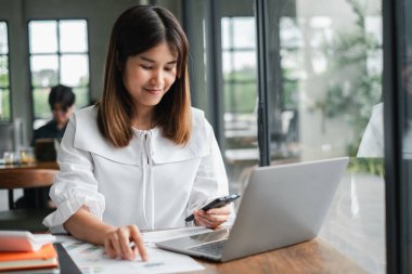 Beyaz bluz giyen genç bir kadın laptopuyla ve akıllı telefonuyla modern bir ofis masasında çalışıyor ve görevlerine odaklanıyor..