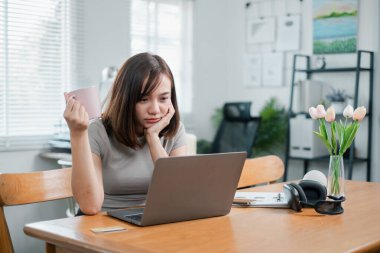 Evinden çalışan genç bir kadın, ahşap bir masada dizüstü bilgisayarı ve kahve fincanı ile parlak, modern bir oturma odasında oturuyor..