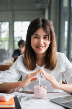Genç bir kadın, elinde bir kumbara tutarken gülümsüyor. Finansal planlamayı ve tasarruf yapmayı simgeliyor..