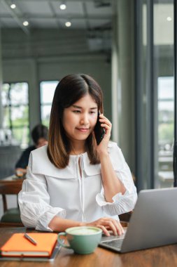 Laptopuyla çalışan ve modern bir kafe ortamında telefonla konuşan genç bir kadın..