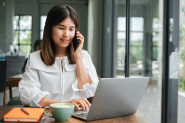 Dizüstü bilgisayarı üzerinde çalışarak ve modern ofis ortamında telefonla konuşarak birden fazla iş yapan genç profesyonel bir kadın..