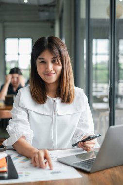 Beyaz bluzlu, kendine güvenen genç bir kadın dizüstü bilgisayarda çalışıyor ve modern ofis ortamında belgeleri inceliyor..