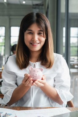 Beyaz bluzlu neşeli bir kadın elinde pembe bir kumbara, geniş pencereli, parlak, modern bir ofiste oturuyor..