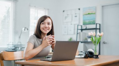 Parlak ve modern bir ofis ortamında dizüstü bilgisayarla çalışırken elinde kahve fincanı tutan gülümseyen genç bir kadın..