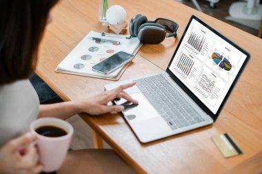 İş kadını finansal veri analizi üzerinde çalışıyor. Laptop, kahve ve belgelerle modern ofis ortamında ahşap bir masada..