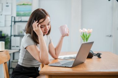 Evinden çalışan genç bir kadın, bir dizüstü bilgisayar ve kahve bardağıyla ahşap bir masada oturuyor, modern bir ofis ortamında..