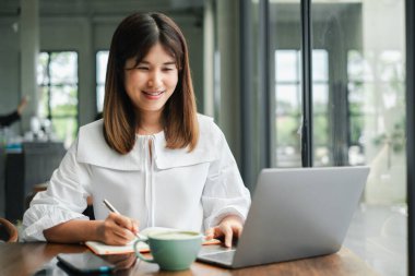 Beyaz bluzlu genç bir kadın dizüstü bilgisayarı üzerinde çalışıyor ve büyük pencereleri ve doğal ışığı olan modern bir kafede notlar alıyor..