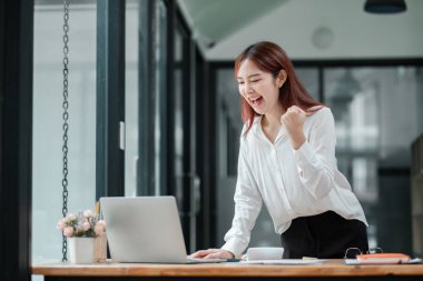 Genç profesyonel bir kadın, modern ofis ortamında dizüstü bilgisayarı üzerinde çalışırken başarılı bir başarıyı kutluyor..