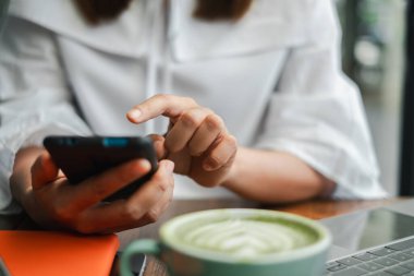 Samimi bir kafede akıllı telefon kullanan bir kadının yakın plan fotoğrafı. Masasında bir fincan kibrit latte ve dizüstü bilgisayar var..