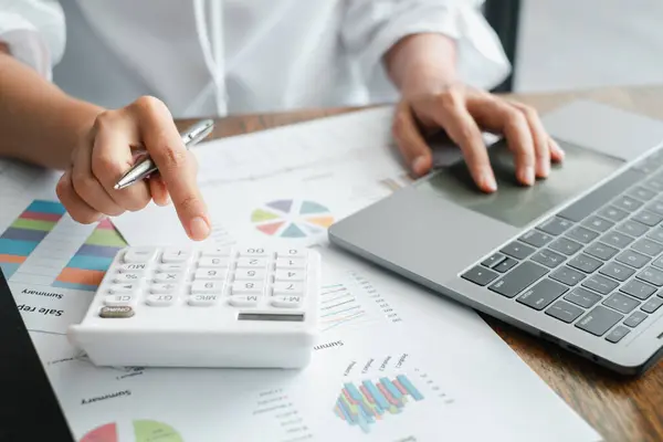 Bir ofis ortamında finansal verileri analiz etmek ve grafikleri gözden geçirmek için hesap makinesi ve dizüstü bilgisayar kullanan bir iş adamının yakın plan fotoğrafı..