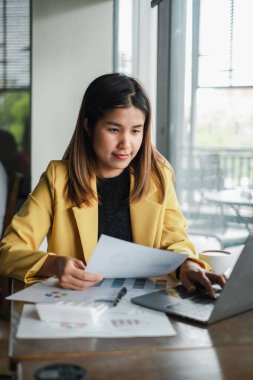 Sarı ceketli genç bir kadın, modern bir ofis ortamında masa başında evrak ve çizelgelerle çalışıyor..