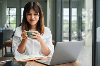 Genç bir kadın elinde bir fincan kahve, modern bir kafede çalışırken gülümsüyor..