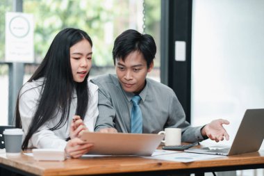 Modern bir ofiste birlikte çalışan iki genç profesyonel, belgeleri tartışıyor ve dizüstü bilgisayar kullanıyor..