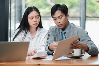 Modern ofis ortamında bir iş projesinde birlikte çalışan iki genç profesyonel, belgeleri gözden geçiriyorlar ve bir dizüstü bilgisayar kullanıyorlar..