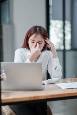 Beyaz gömlekli bir iş kadını, masasında dizüstü bilgisayarı ve modern bir ofis ortamında dokümanlarıyla çalışırken strese giriyor..