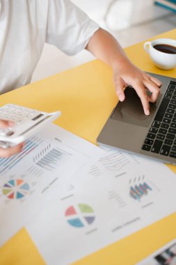Bir dizüstü bilgisayar ve hesap makinesi üzerinde çalışan, sarı bir masada mali tabloları olan, bir fincan kahveyle birlikte bir kişinin yakın çekimi..