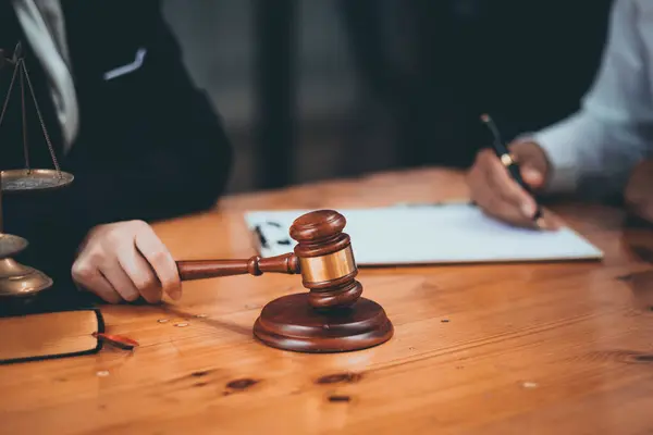 Arkaplanda yasal belgeler ve adalet terazisi olan ahşap bir masanın üzerindeki yargıç tokmağının yakın çekimi..