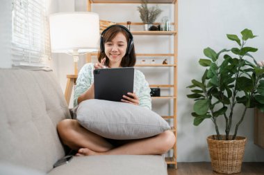 Kanepede oturan, tablet kullanan ve modern bir oturma odasında şık dekorlu kulaklık takan genç bir kadın..