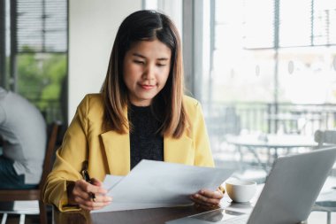 Masasında doküman ve dizüstü bilgisayarla çalışan sarı ceketli genç bir kadın modern ofis ortamındaki görevlerine odaklanmış..