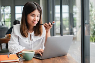 Modern bir kafede uzaktan çalışan genç bir kadın, ses komutları için dizüstü bilgisayar ve akıllı telefon kullanıyor, masada bir fincan kahve ve bir not defteri var..