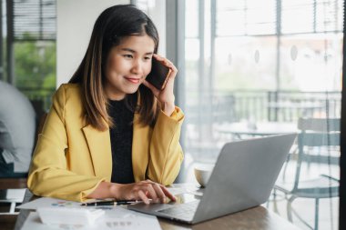 Sarı ceketli genç bir kadın, çağdaş bir ofis ortamında telefonla konuşurken dizüstü bilgisayarında çalışıyor..