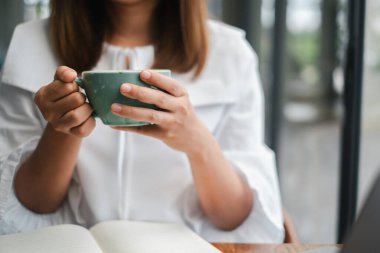 Elinde yeşil kahve fincanı olan bir kadın, açık bir kitapla masada oturuyor, modern bir kafede huzurlu bir anın tadını çıkarıyor..