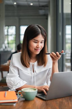 Modern bir kafede dizüstü bilgisayarı üzerinde çalışan beyaz bluzlu genç bir kadın elinde bir kalem, elinde gülücükler, masada kahve ve bir defterle..