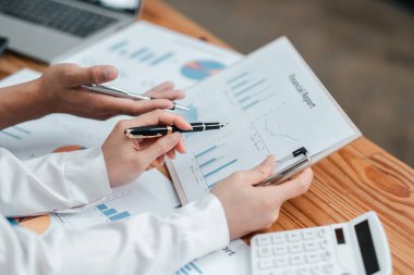 Kalemleri tutan ellerin yakın plan çekimi ve ahşap bir tablodaki tablolar ve grafiklerle mali raporları tartışmak.