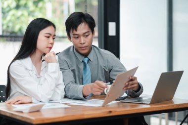 Two professionals engaged in a business discussion, reviewing documents and using a laptop in a modern office environment. clipart