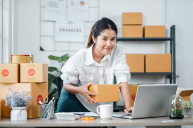 Çağdaş bir ofis ortamında dizüstü bilgisayarı üzerinde çalışırken teslimat için paketler hazırlayan genç bir iş kadını..