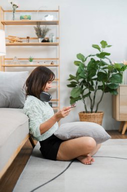 Genç bir kadın yerde oturuyor, akıllı telefonunu kullanıyor ve rahat, modern bir oturma odasında kulaklık takıyor bitkilerle ve ahşap mobilyalarla..