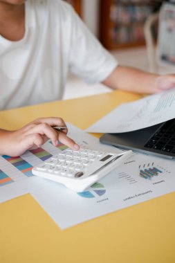Finansal analiz için hesap makinesi ve dizüstü bilgisayar kullanan, sarı bir masanın üzerindeki grafikler ve çizelgeler ile çevrili bir kişinin yakın plan görüntüsü..