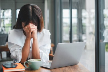 Bir kadın evde çalışırken stresli görünüyor, modern bir ofis ortamında dizüstü bilgisayar ve kahveyle bir masada oturuyor..