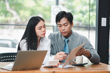 Modern bir ofiste çalışan iki genç profesyonel, belgeleri tartışıyor ve bir dizüstü bilgisayar kullanıyorlar..