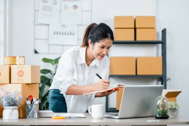 Genç bir kadın dizüstü bilgisayarda çalışıyor ve elektronik ticareti ve küçük işletmeleri temsil eden kargo kutularıyla modern bir ev ofisinde notlar alıyor..